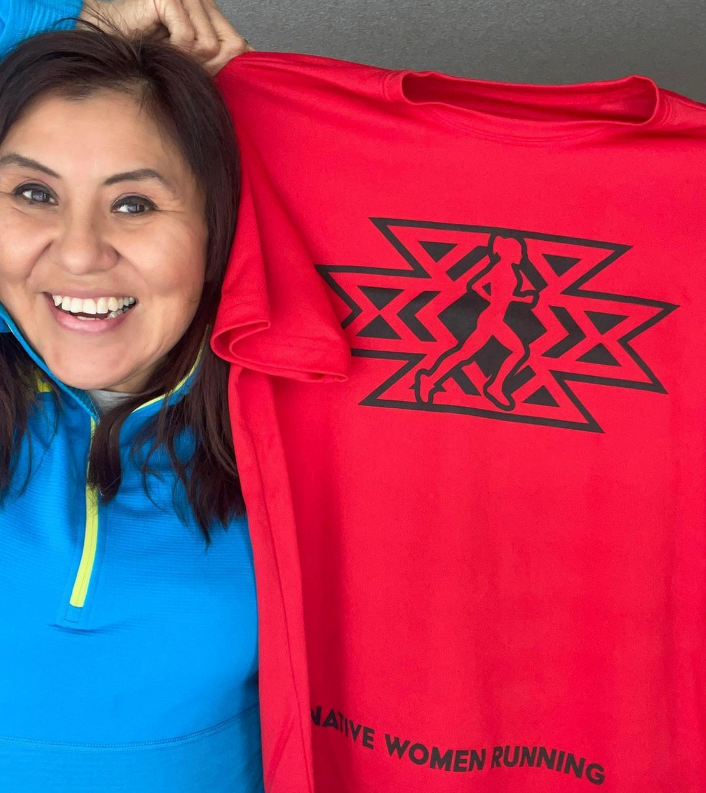 A smiling woman (Verna Volker) holds a red tshirt that reads "Native Women Running."