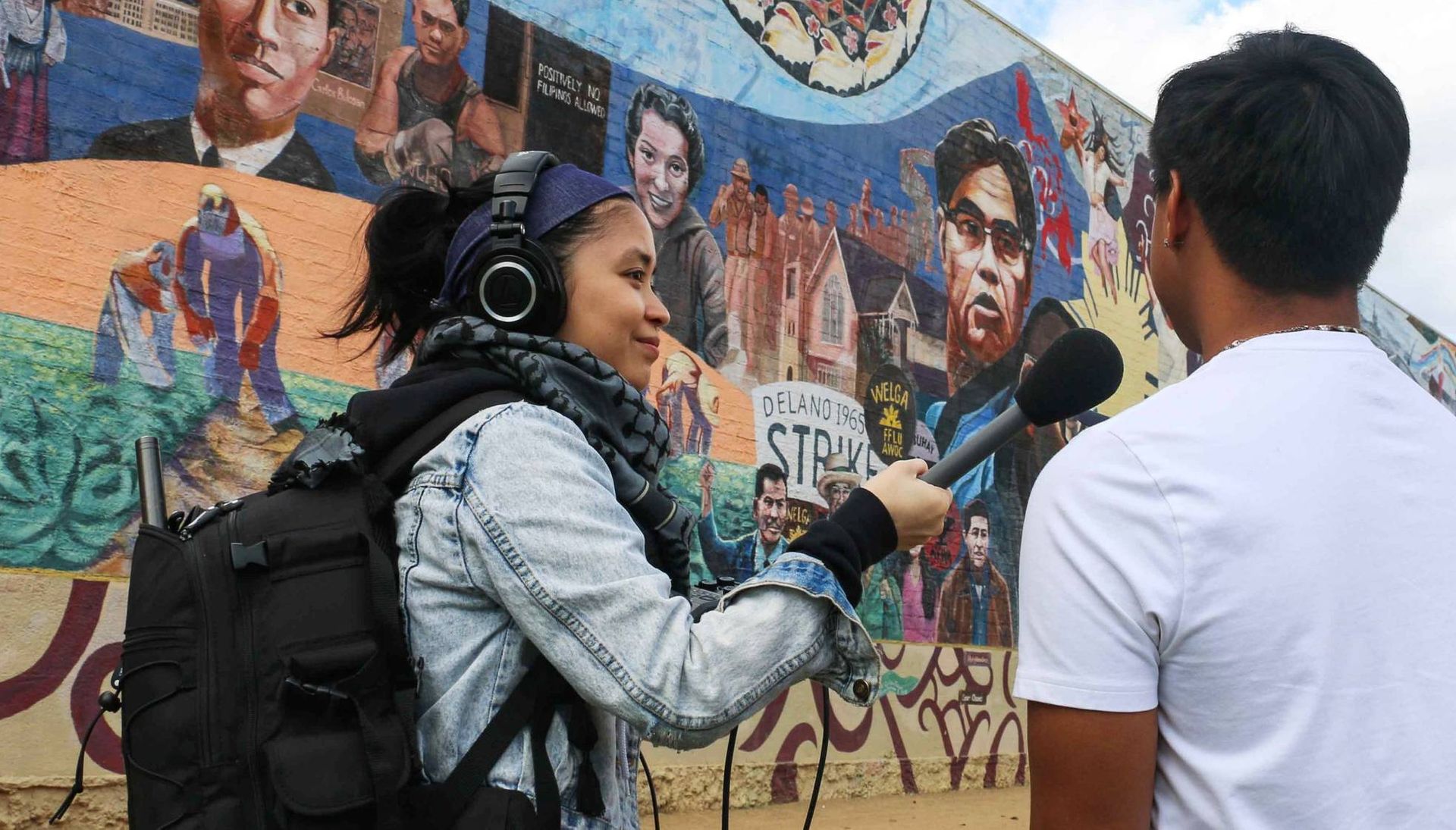 A young woman interviews a person in front of a mural