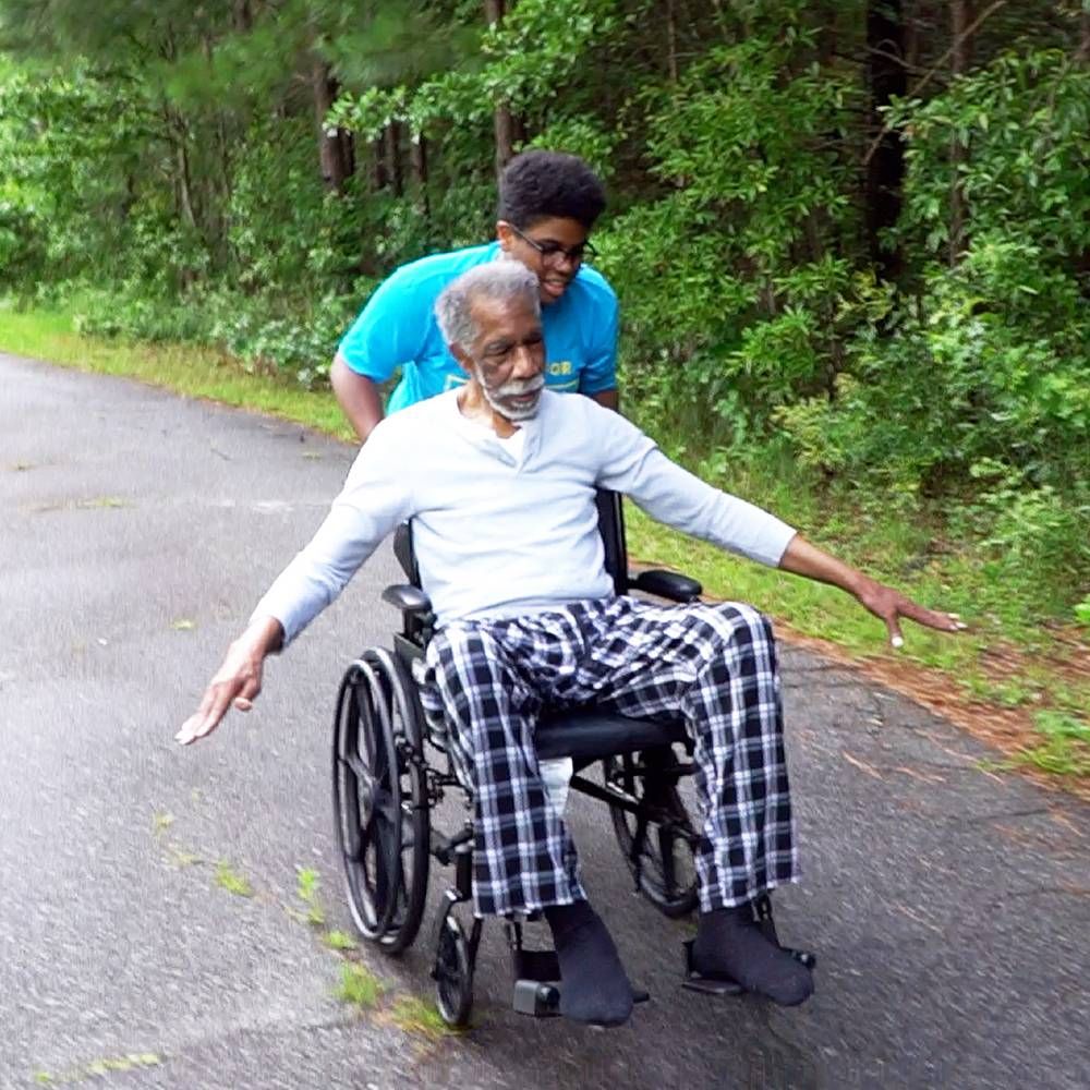 A young Black man pushes his grandfather in a wheelchair. Rewire PBS Love Caregiver Lumiere Rostick