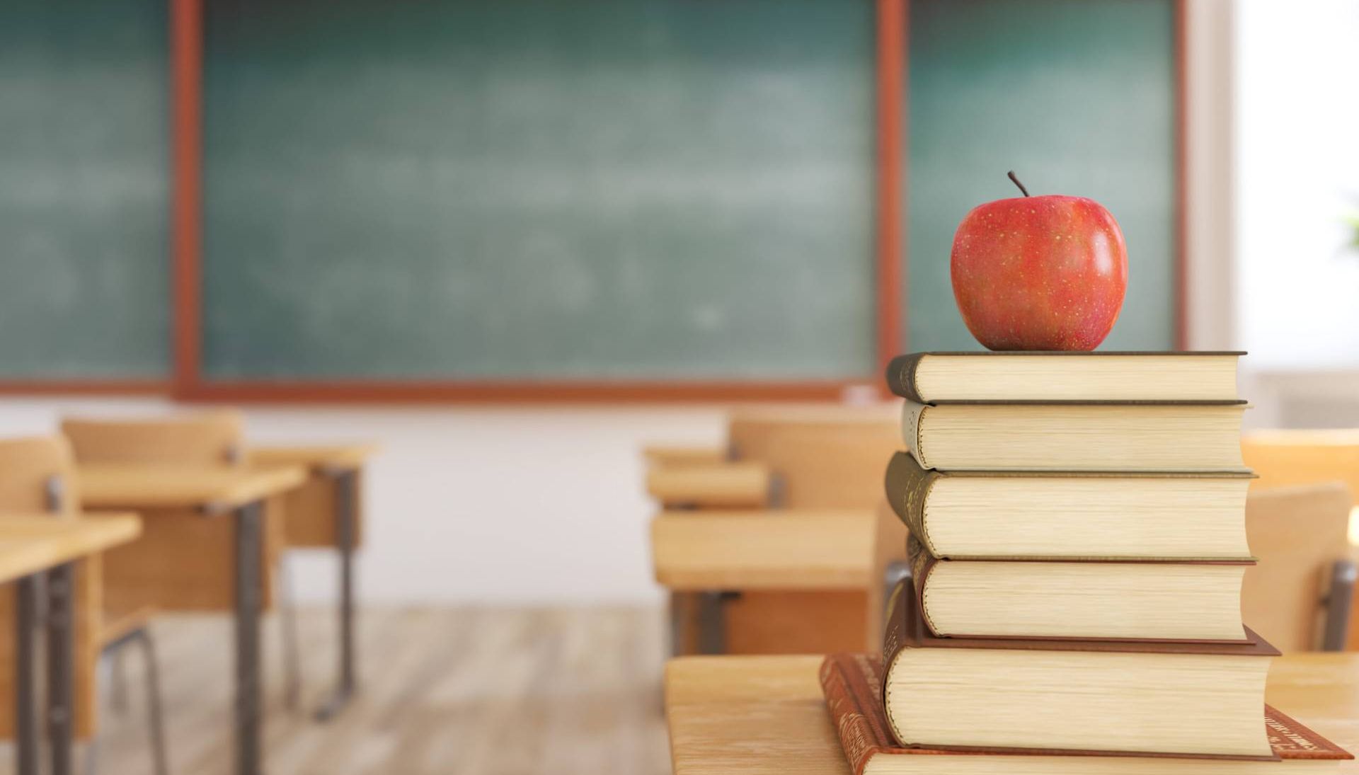 Photo of one red apple on top of books in an empty school classroom.