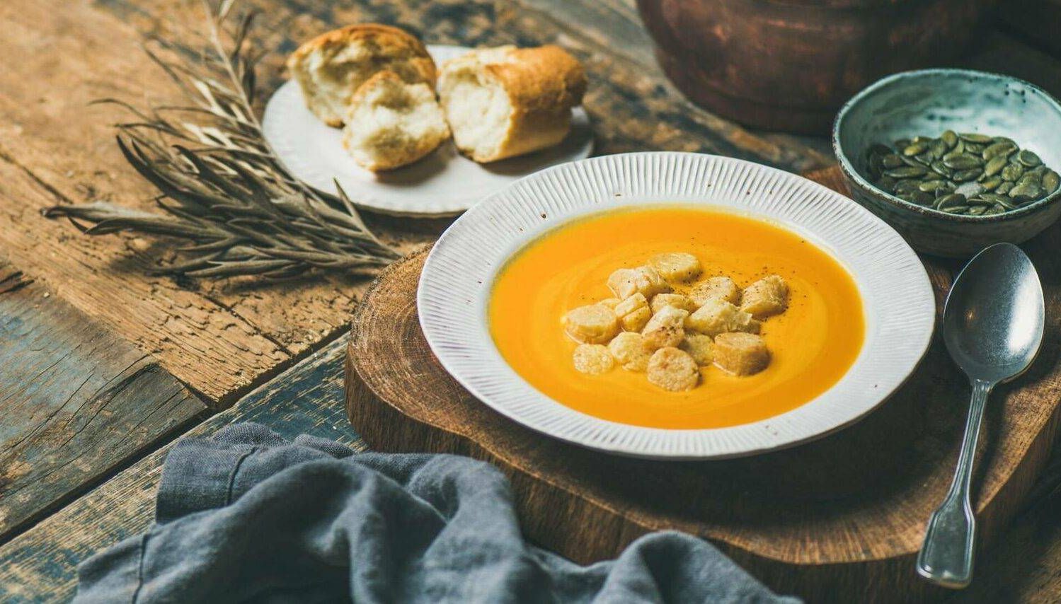 Fall warming pumpkin cream soup with croutons and seeds on board over rustic wooden background, copy space, wide composition. Autumn vegetarian, vegan, healthy comfort food concept. Rewire PBS Living Comfort Foods