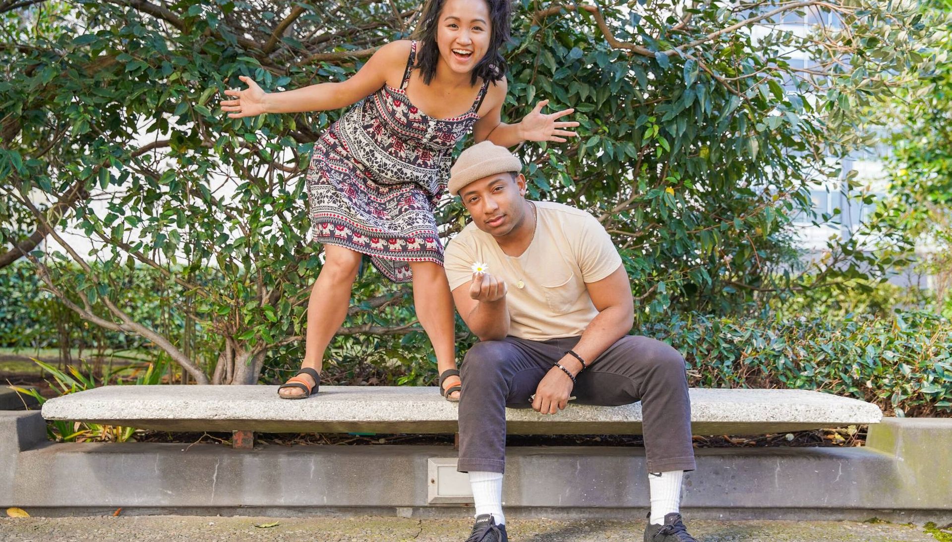 Photograph of two young people on a bench outdoors, one man sitting and holding a flower and one woman standing with her arms spread in front of a tree