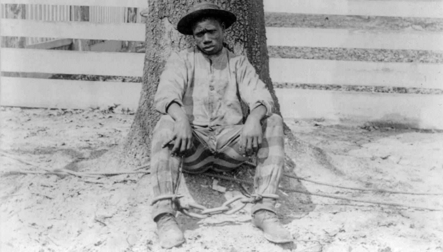 Black and white image of a Black man sitting against a tree, wearing a prison uniform and shackles. Slavery by Another Name, PBS, Rewire, our future, 